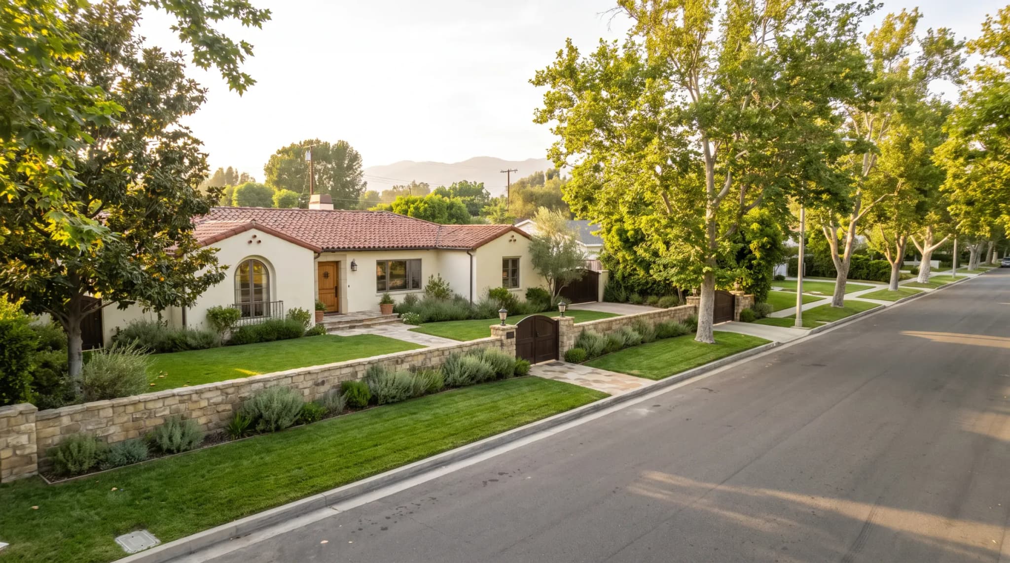 Encino Mediterranean revival home with cream stucco, terracotta tile roof, and a deep front lawn behind a low stone wall with wooden gate, framed by mature deciduous trees and distant mountain ridgelines.